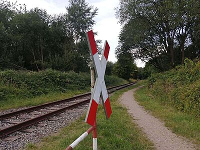 Das Foto zeigt im Vordergrund ein Andreaskreuz für einen unbeschrankten Bahnübergang, im Hintergrund die auf Gleisbett verlaufende Schiene. 